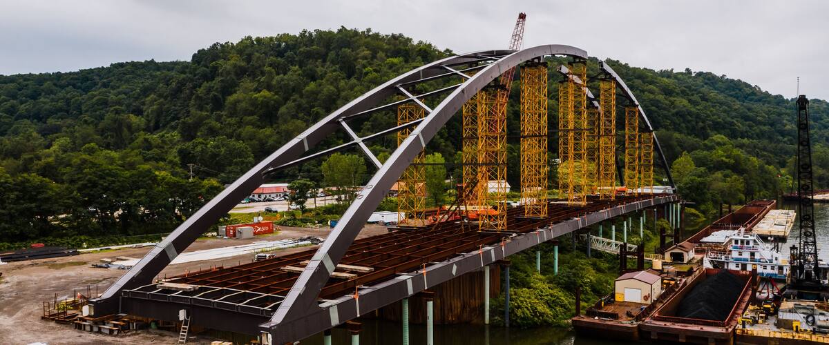 A large steel bridge with a curved arch spanning a river, surrounded by dense forest and industrial structures.