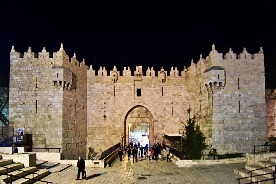 A historic stone gate with battlements, a central archway, and a crowd of people approaching it.