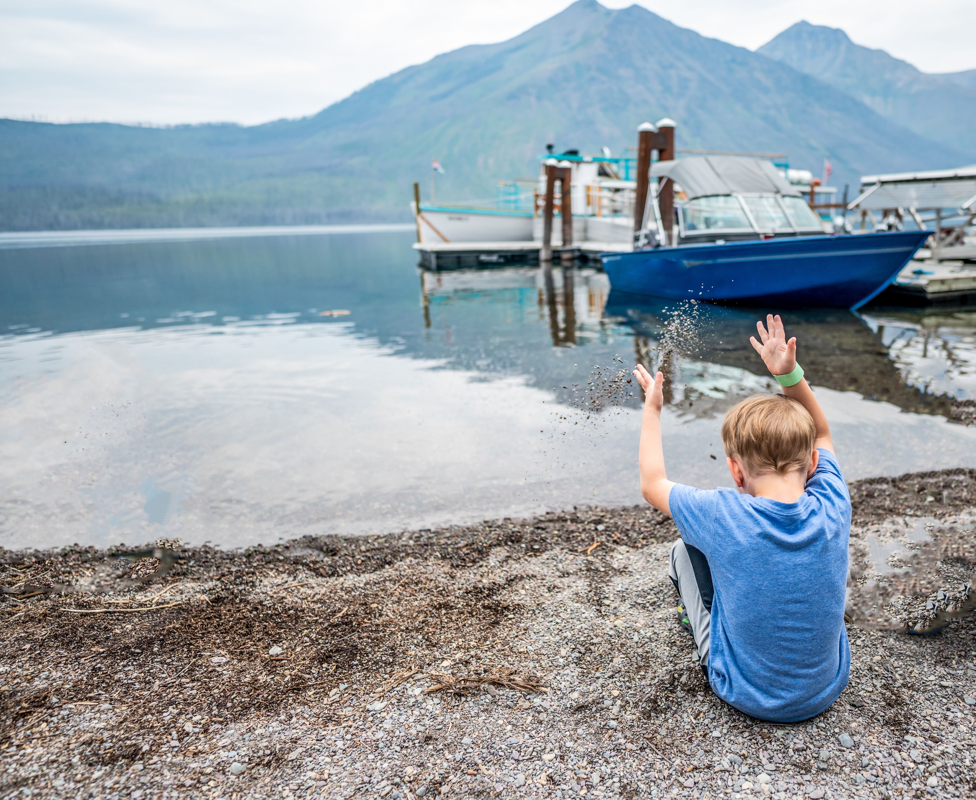Un niño jugando junto a un lago con barcos amarrados al fondo y montañas a lo lejos.