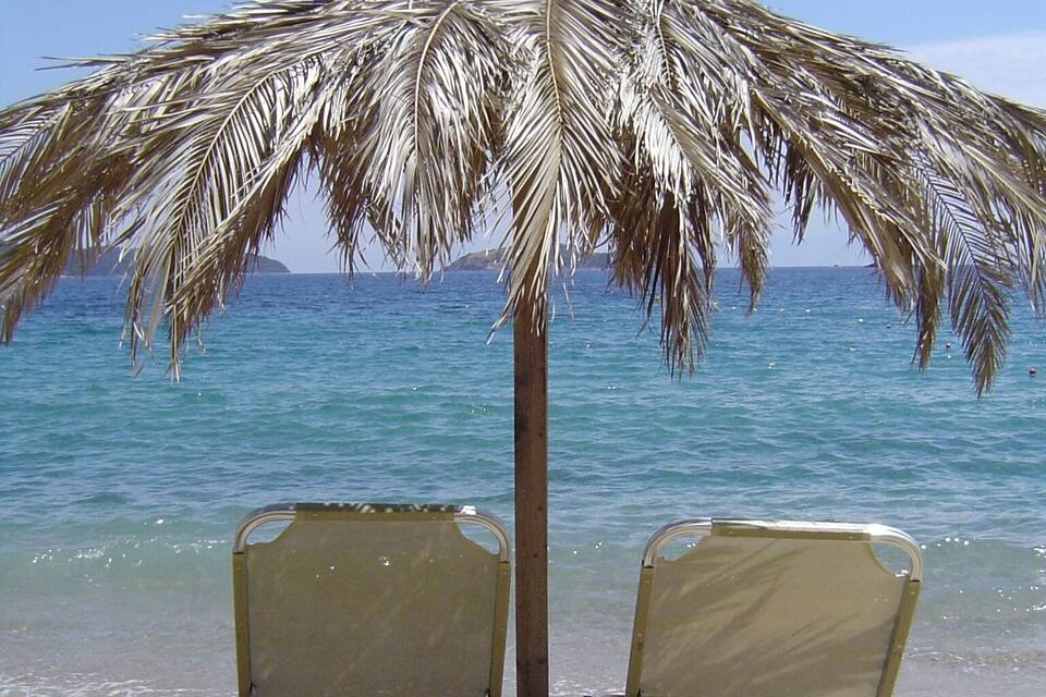 Two beach chairs under a palm tree on a sandy beach with a clear blue ocean and distant land in the background.