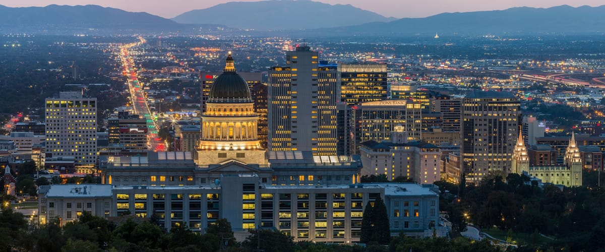 A cityscape at dusk with illuminated buildings, a prominent dome structure, and a distant mountain range.