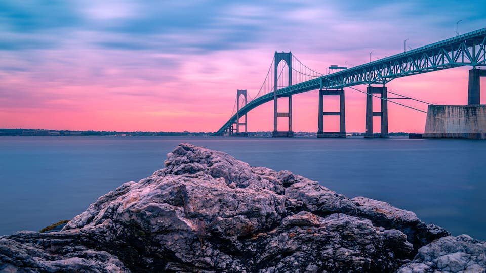 A large suspension bridge over a calm body of water at dusk with a rocky foreground.