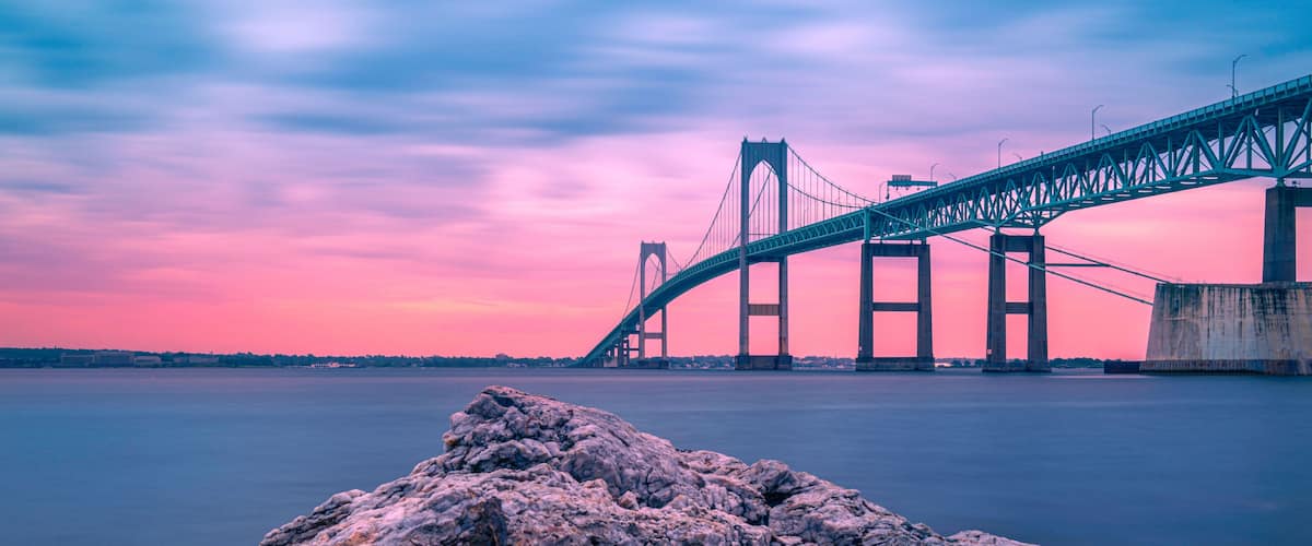 A large suspension bridge over a calm body of water at dusk with a rocky foreground.