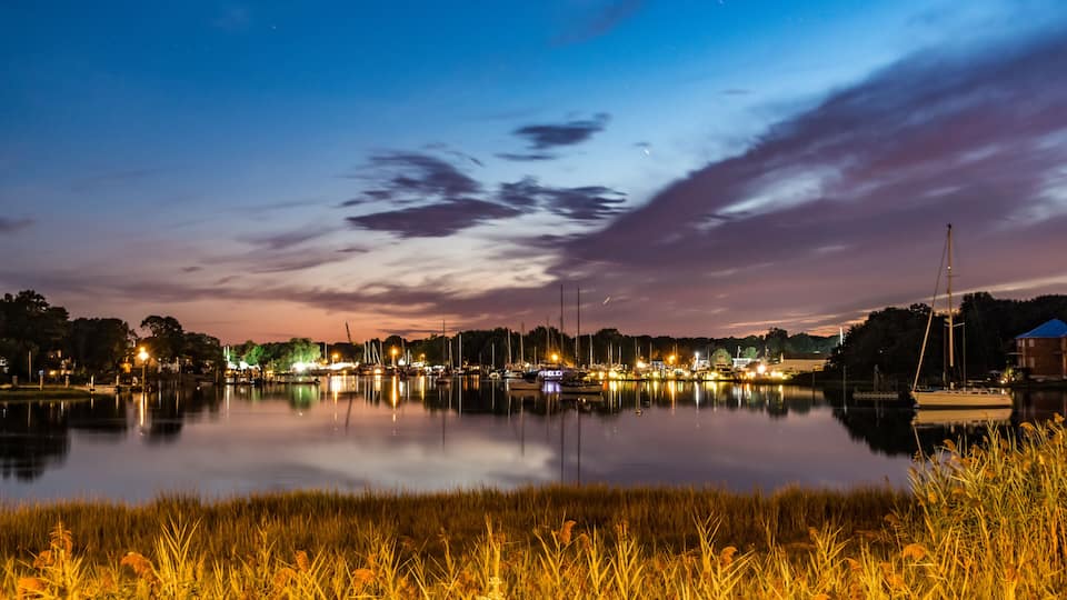 A calm lake at dusk with boats and a sailboat docked, surrounded by grassy banks and a distant city skyline.