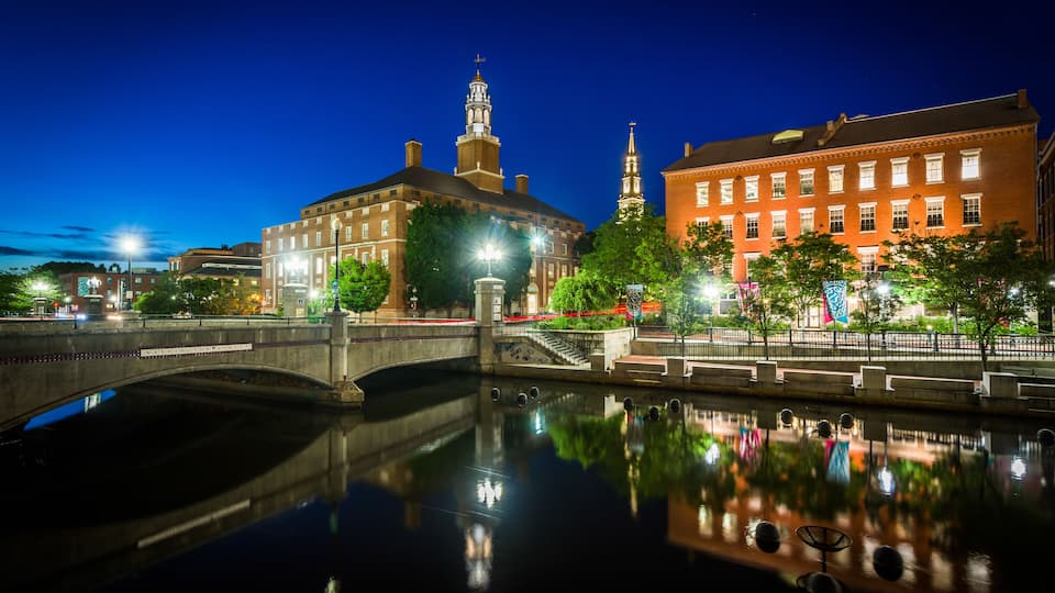A nighttime cityscape with a bridge over a calm river, reflecting buildings and streetlights.