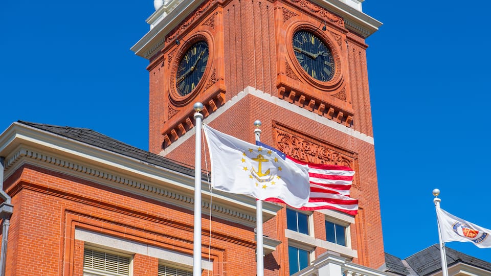 A red brick clock tower with two large clocks, a white flag with an anchor emblem, and the American flag.