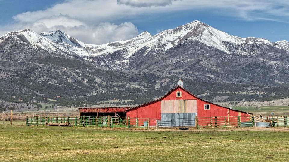 A red barn with a green fence in a grassy field, with snow-capped mountains in the background.