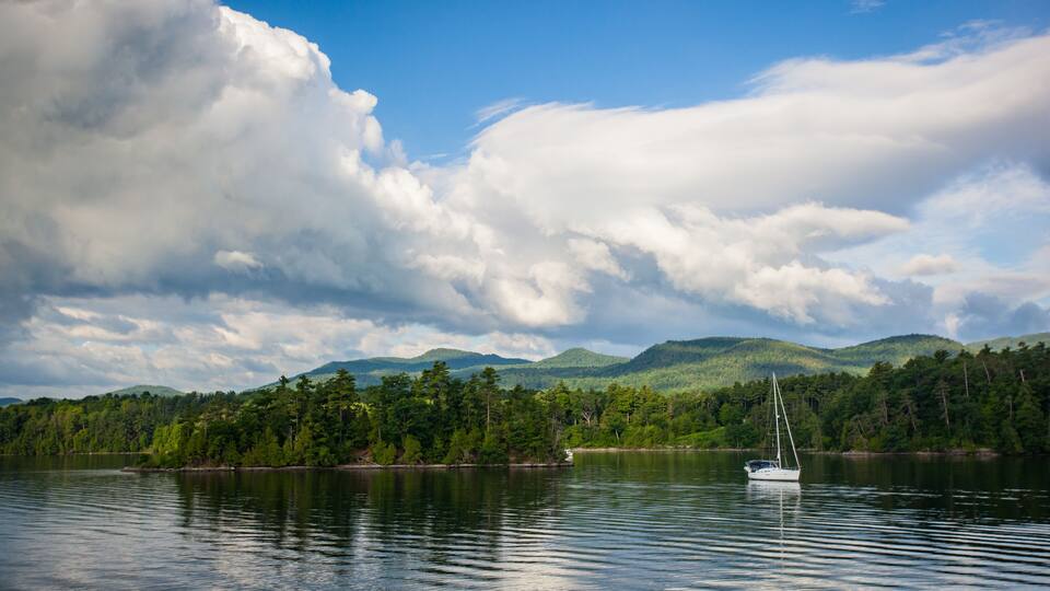 A calm lake with a sailboat, surrounded by lush green hills and a partly cloudy sky.