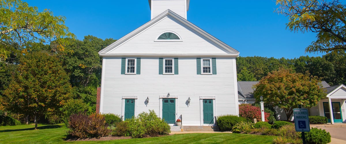 A white church with a steeple and green shutters, surrounded by trees and a clear blue sky.