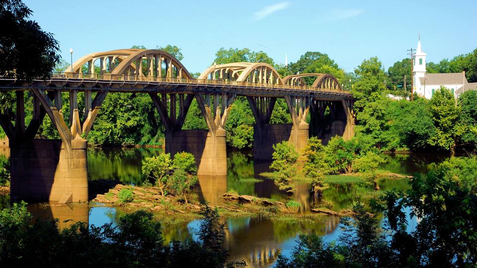 A steel bridge with arches over a river, surrounded by lush greenery and a church in the background.
