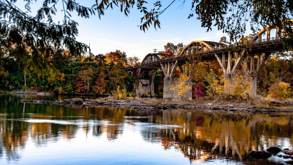 A bridge over a calm river with autumn foliage and a church in the background.