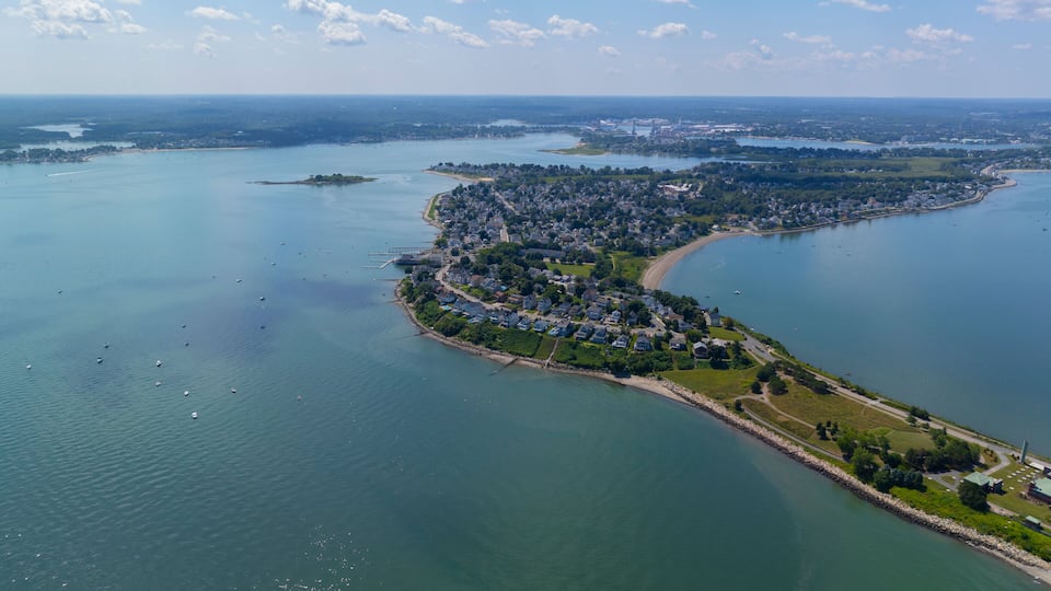 An aerial view of a coastal town with a curved shoreline, a marina, and a bridge connecting to the mainland.