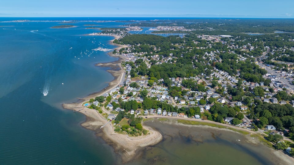 An aerial view of a coastal town with a beach, marina, and dense residential area.