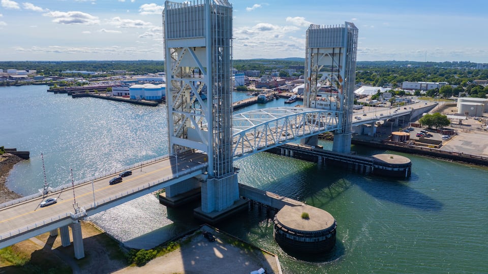 A large steel bridge spanning a river with industrial buildings and a parking lot in the foreground.