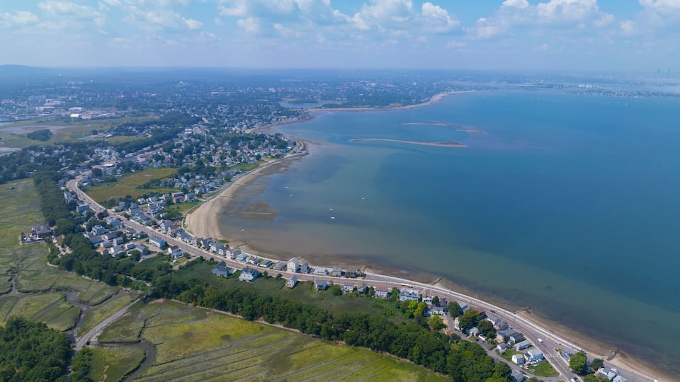 An aerial view of a coastal town with a curved road along the beach, a large body of water, and surrounding green fields.