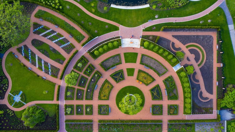 An aerial view of a landscaped park with winding paths, a central circular garden, and a fountain.