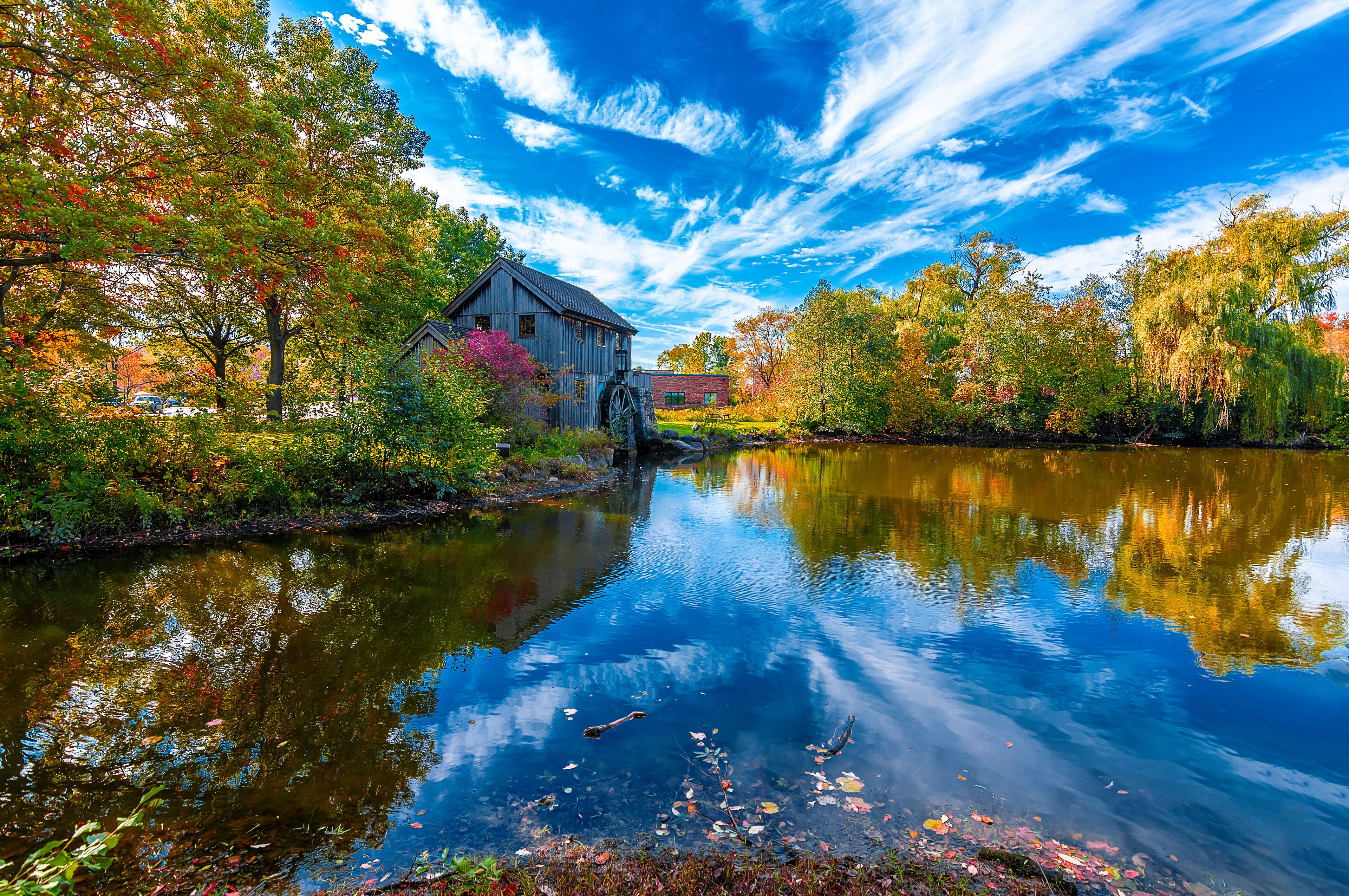 A rustic mill by a calm river, surrounded by autumn foliage and a clear blue sky.