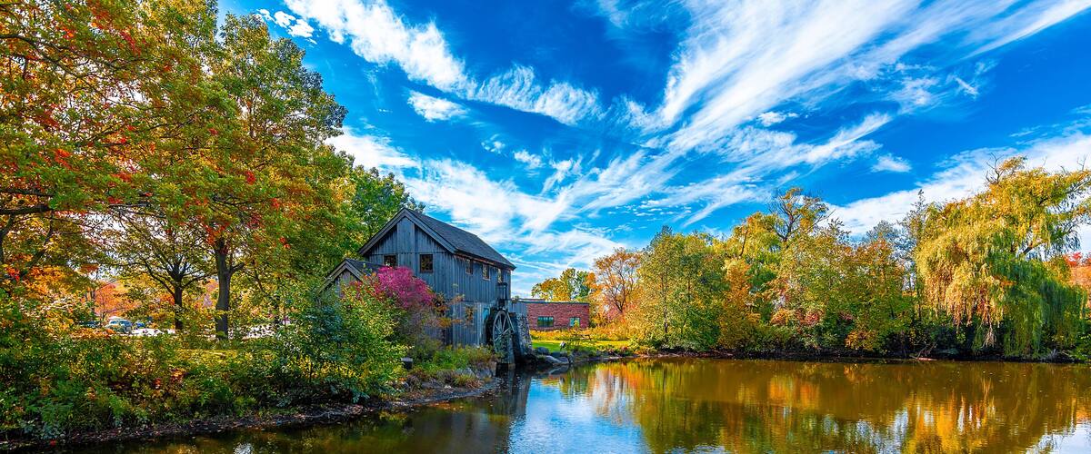 A rustic mill by a calm river, surrounded by autumn foliage and a clear blue sky.