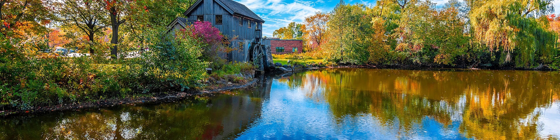 A rustic mill by a calm river, surrounded by autumn foliage and a clear blue sky.