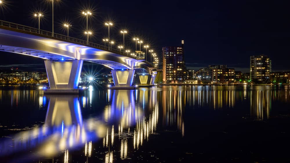 Ein Nachtblick auf eine beleuchtete Brücke, deren Lichter sich im Wasser spiegeln, mit einer Stadtansicht im Hintergrund und einem klaren Himmel.