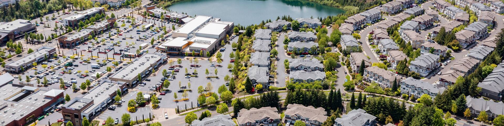 Aerial drone view of Beaverton, Oregon, featuring Progress Lake Park, residential neighborhoods, shopping centers, and parking lots, with tree-lined streets and a mix of suburban and commercial spaces