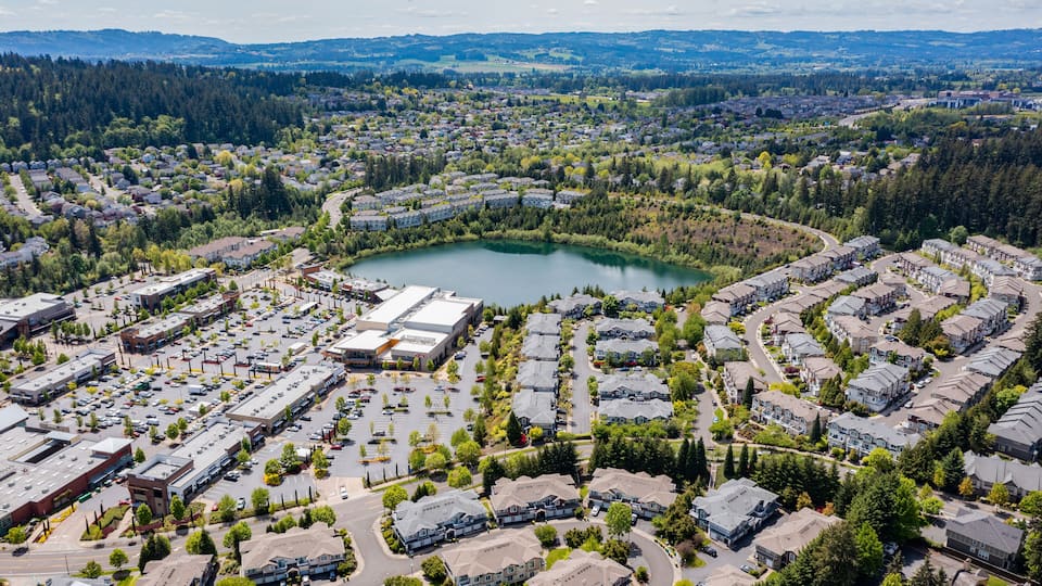 Aerial drone view of Beaverton, Oregon, featuring Progress Lake Park, residential neighborhoods, shopping centers, and parking lots, with tree-lined streets and a mix of suburban and commercial spaces