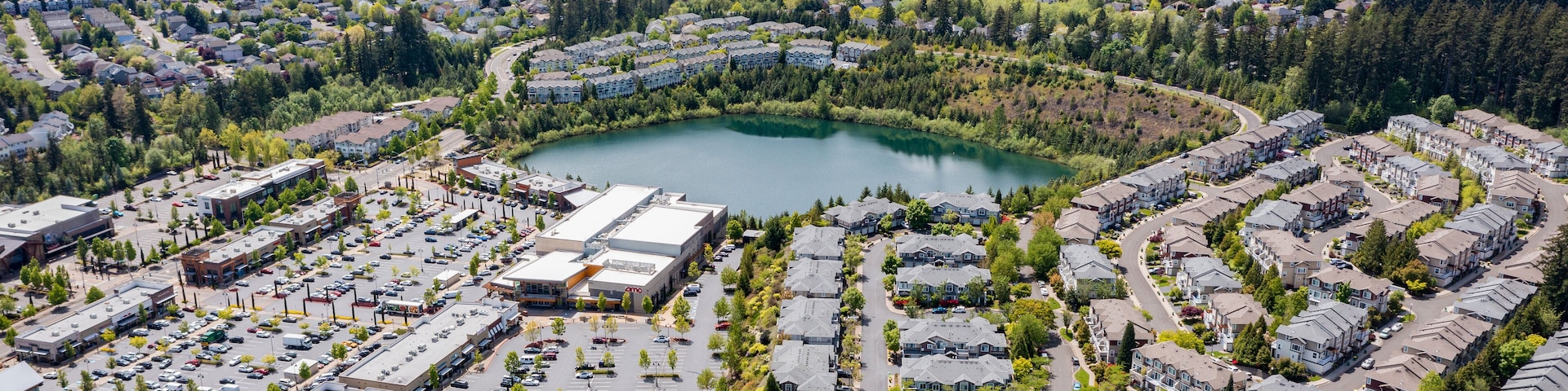 Aerial drone view of Beaverton, Oregon, featuring Progress Lake Park, residential neighborhoods, shopping centers, and parking lots, with tree-lined streets and a mix of suburban and commercial spaces