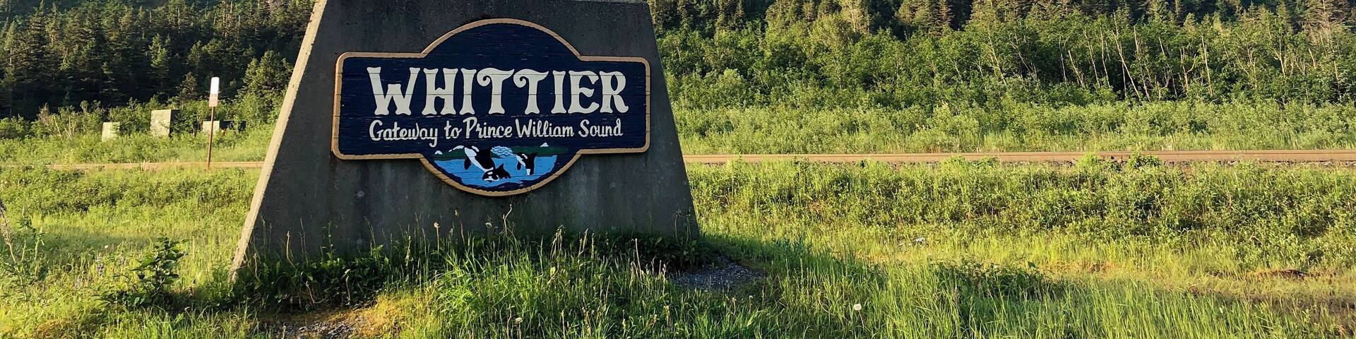 A sign reading "Whittier Gateway to Prince William Sound" with a mountainous backdrop.