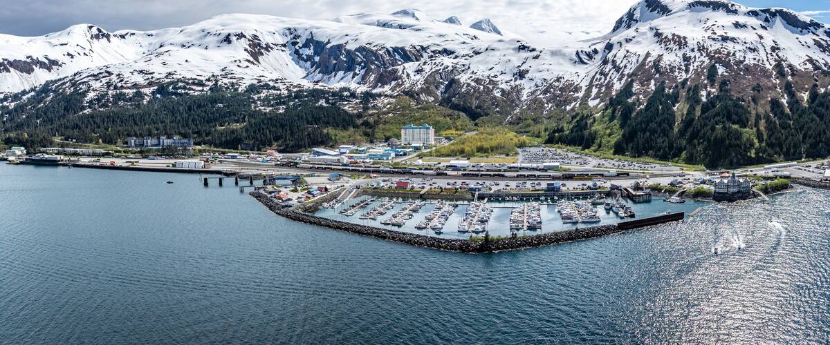 A coastal town with a marina, a large building, and snow-capped mountains in the background.