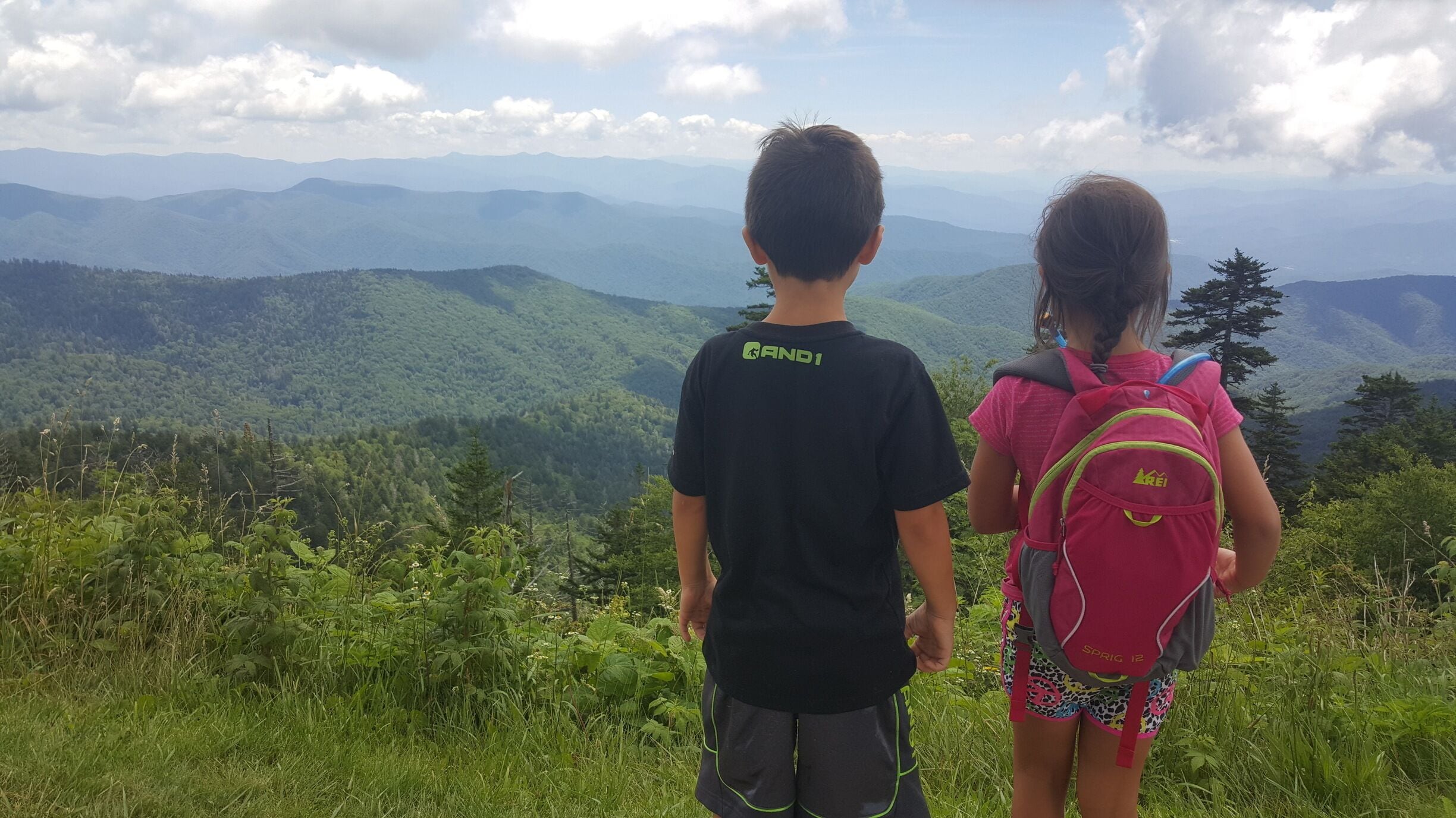 Two children standing on a grassy hill overlooking a mountainous landscape.