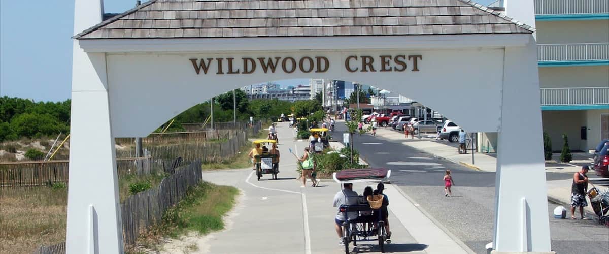 A pedestrian pathway leading to a sign reading "Wildwood Crest" with people walking and a beach in the background.
