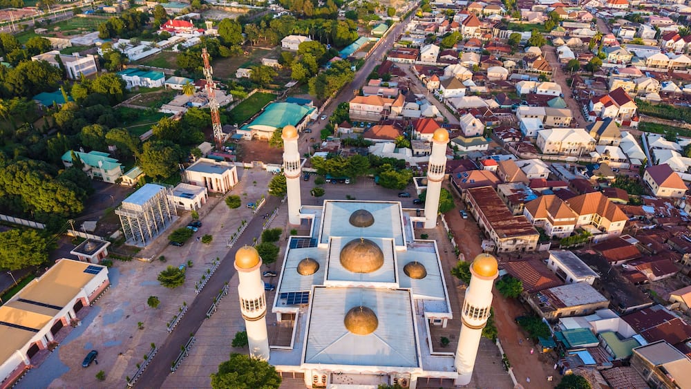 An aerial view of a mosque with four golden domes and two minarets, surrounded by residential buildings and greenery.