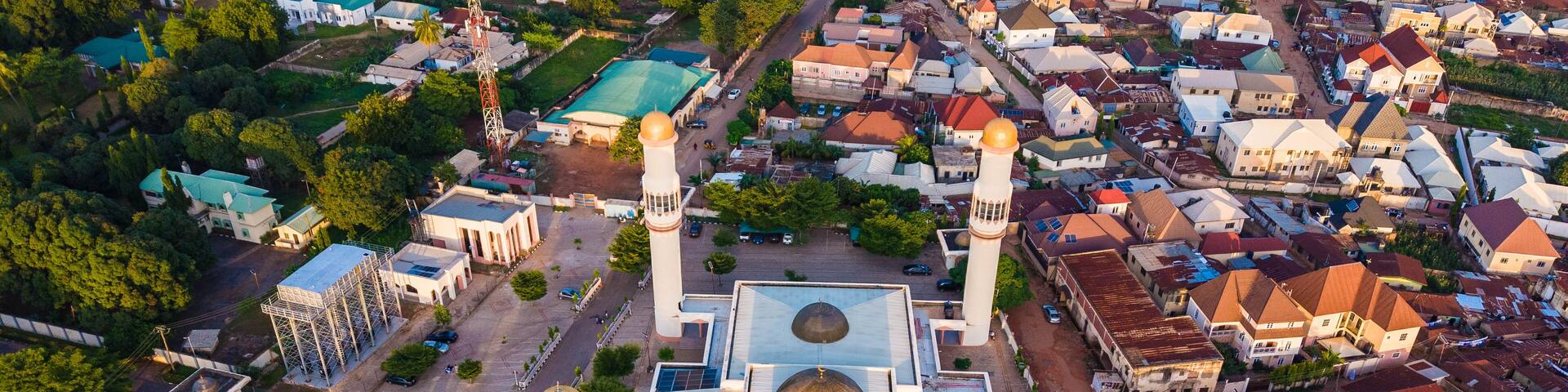 An aerial view of a mosque with four golden domes and two minarets, surrounded by residential buildings and greenery.