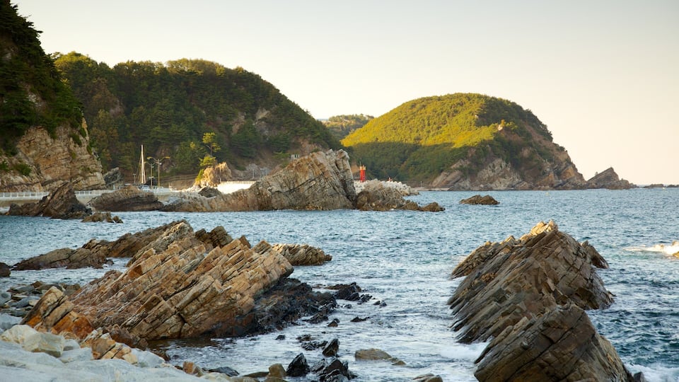 A rocky coastline with waves crashing against the rocks, a lighthouse, and a hill with trees in the background.