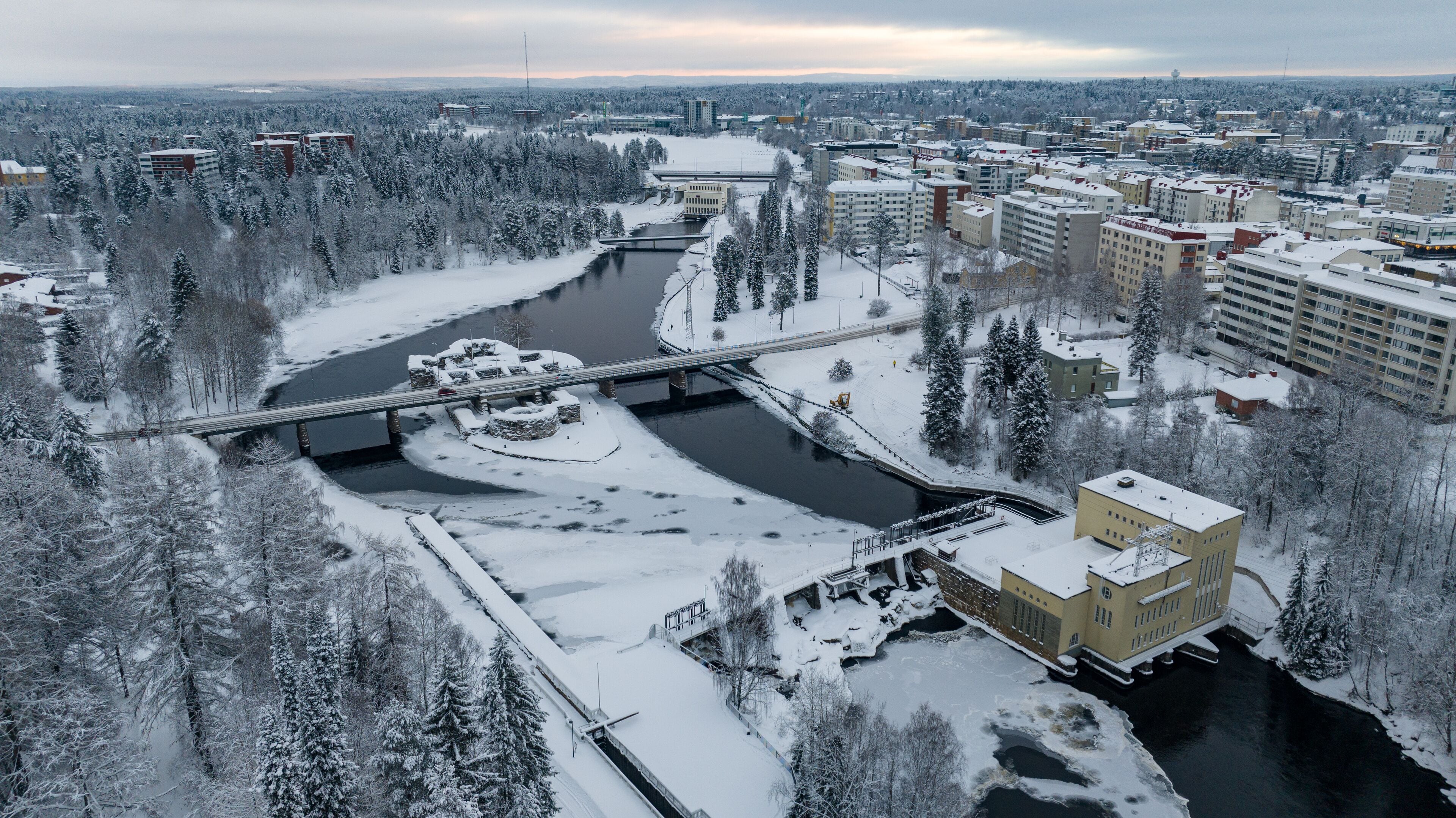 Aerial view of a snow-dusted landscape where the Kajaani river winds through icy patches, meeting a hydro-electric power plant near the bridge, Kajaani, Kainuu, Finland.