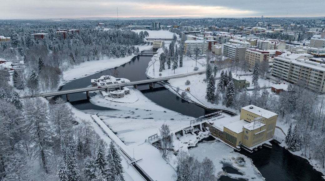 Aerial view of a snow-dusted landscape where the Kajaani river winds through icy patches, meeting a hydro-electric power plant near the bridge, Kajaani, Kainuu, Finland.