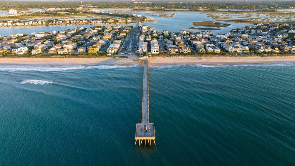 An aerial view of a coastal town with a long pier extending into the ocean, surrounded by residential buildings and a sandy beach.