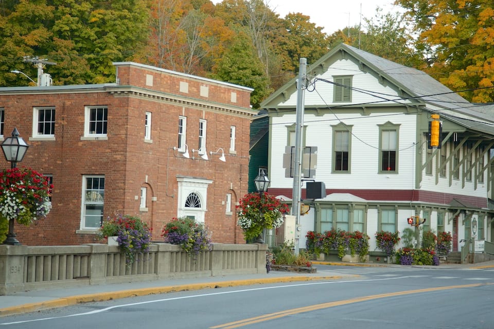 A street with a red brick building and a white building with green shutters. There are flower boxes on the sidewalk.