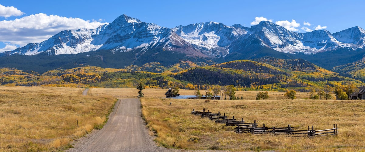 A dirt road leading to a wooden fence, with snow-capped mountains in the background and autumn-colored trees.