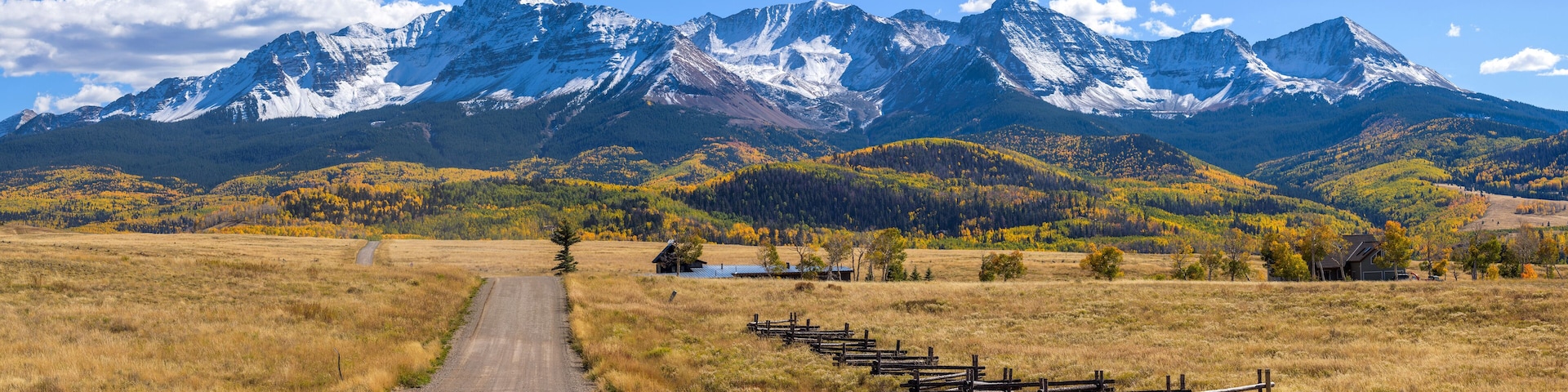 A dirt road leading to a wooden fence, with snow-capped mountains in the background and autumn-colored trees.