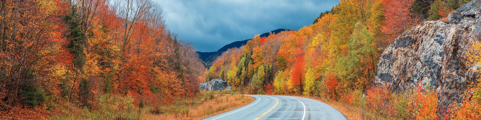 A winding road through a forest with autumn foliage, a rocky cliff on the right, and a mountain in the background.