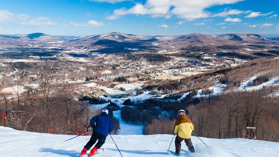 Two skiers on a snowy slope with a valley and mountains in the background.