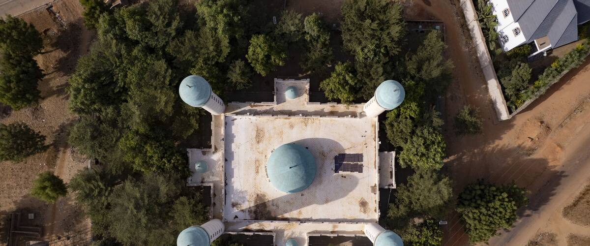 An aerial view of a square building with a central dome and four smaller domes, surrounded by trees and a dirt road.