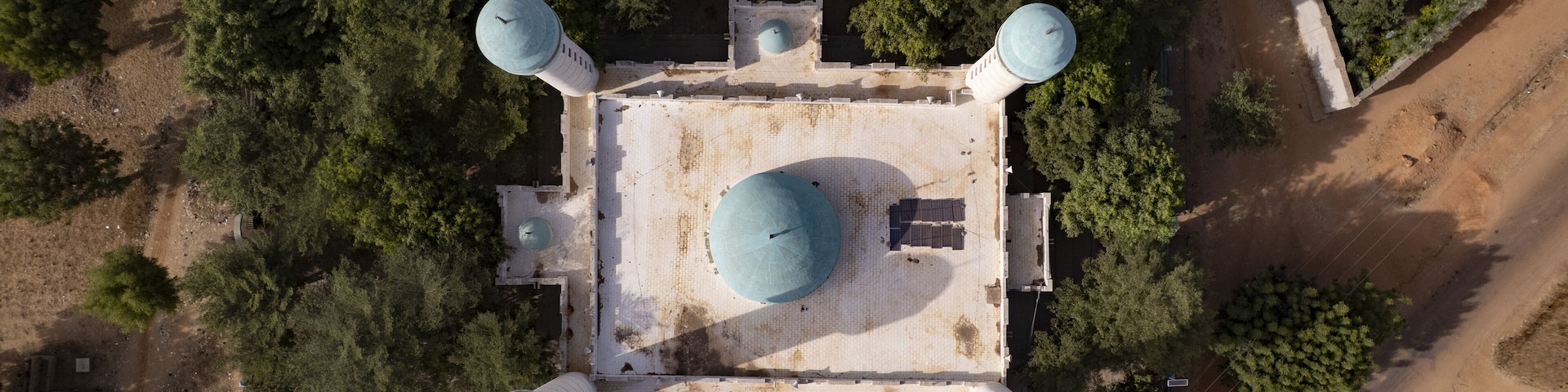 An aerial view of a square building with a central dome and four smaller domes, surrounded by trees and a dirt road.