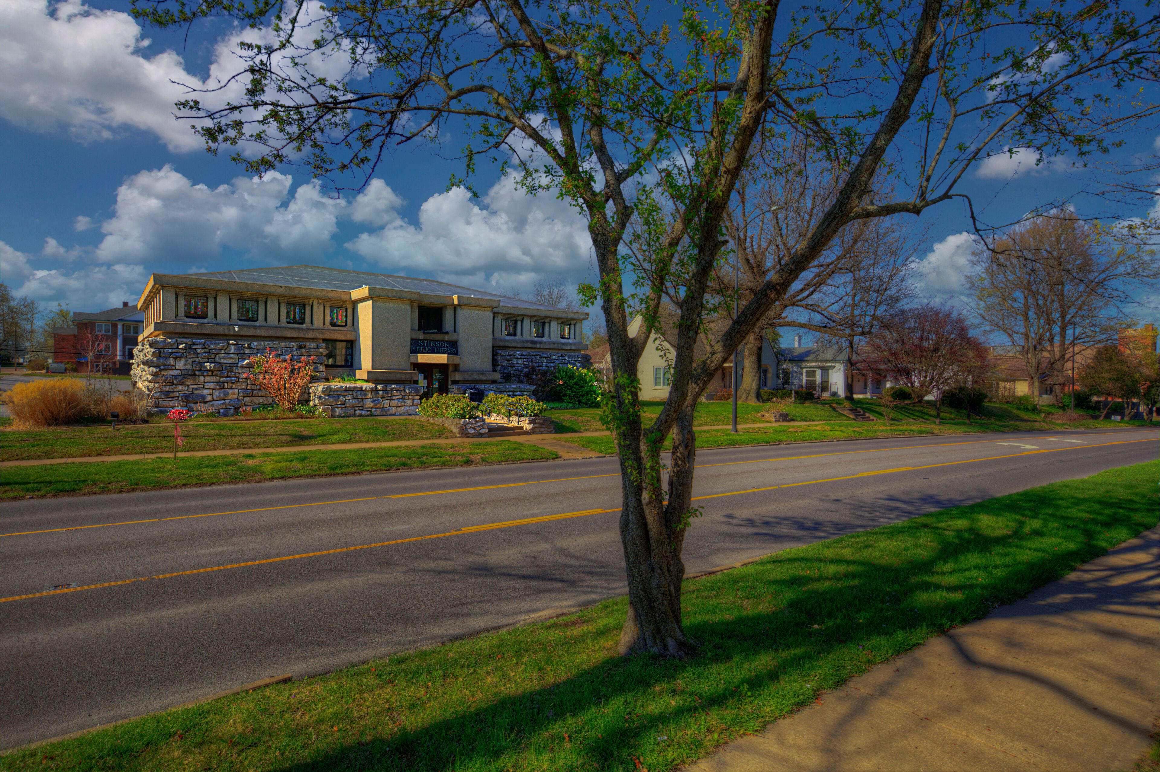 A suburban street with a stone house, a tree, and a clear sky.