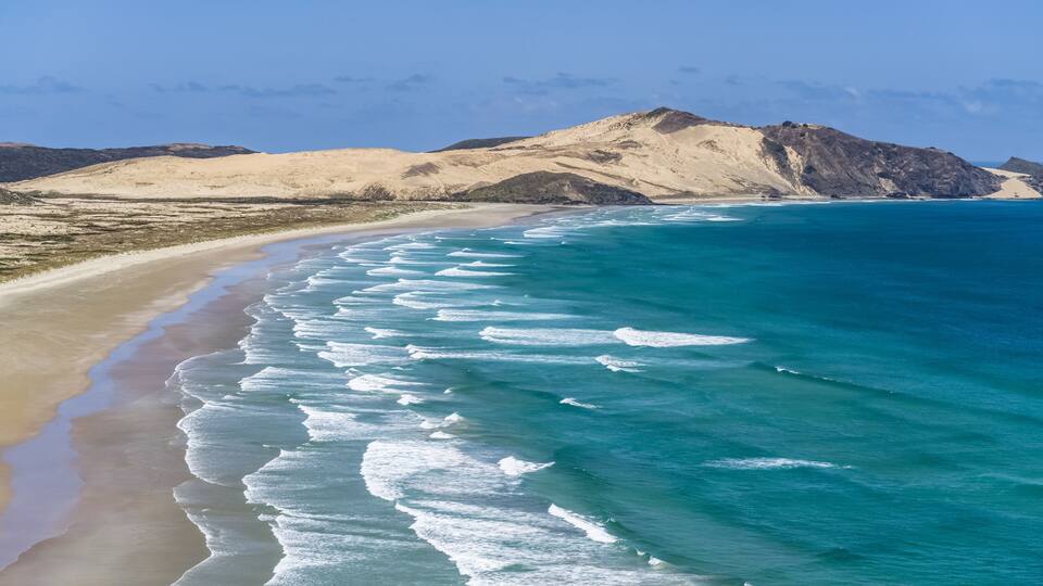 A beach with turquoise waves and a mountainous backdrop.