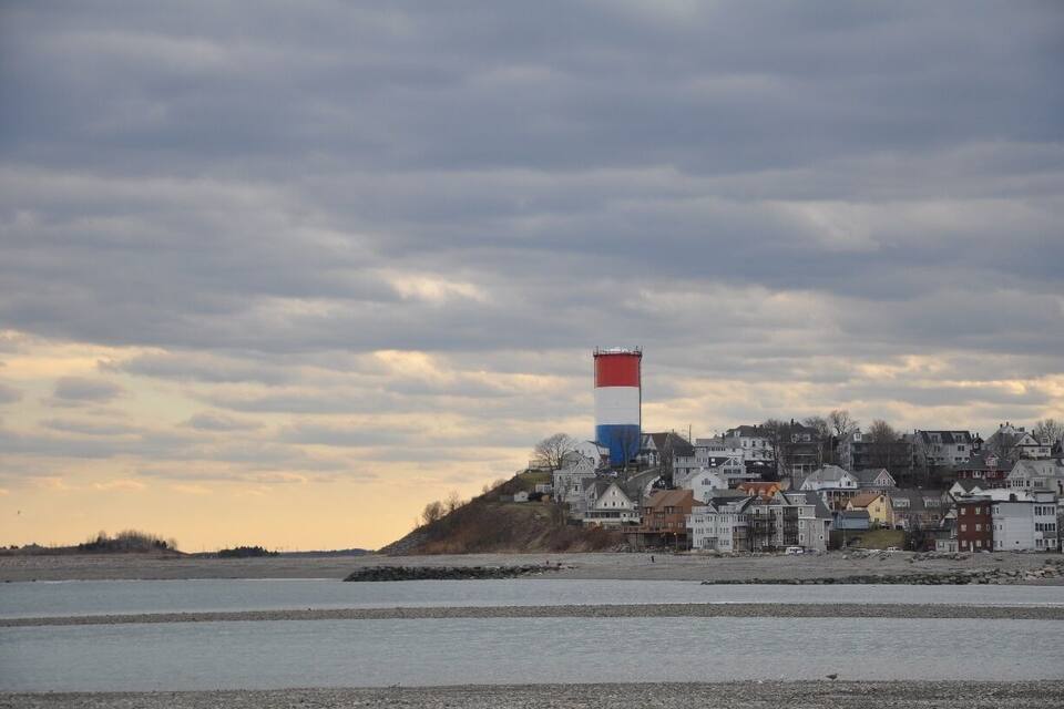 A lighthouse with a red, white, and blue top, a beach, and a town with houses.