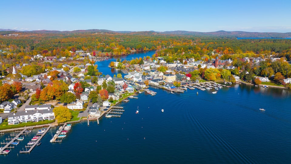An aerial view of a lakeside town with colorful autumn foliage, a church, and a marina with boats.