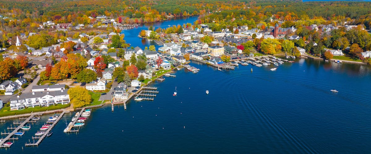 An aerial view of a lakeside town with colorful autumn foliage, a church, and a marina with boats.