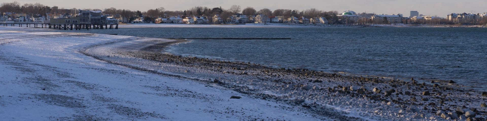 A snow-covered beach with a pier extending into the water, a row of houses in the background, and a clear sky.